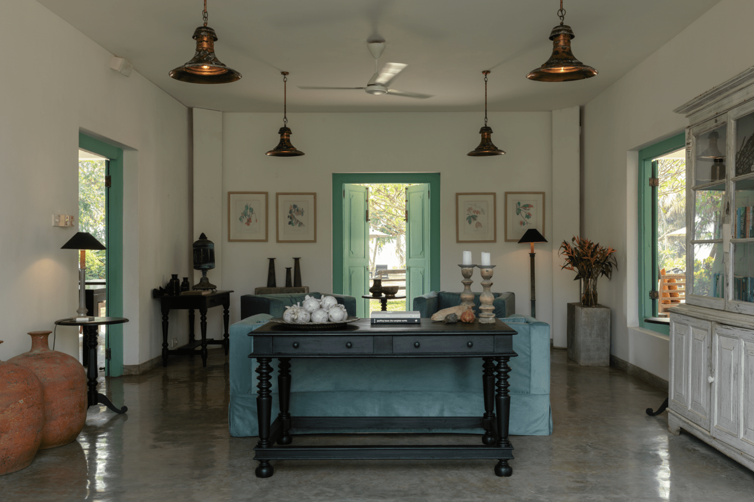 A white living room with a grey polished floor, pendant lamps, a blue sofa and teal doors open to the garden