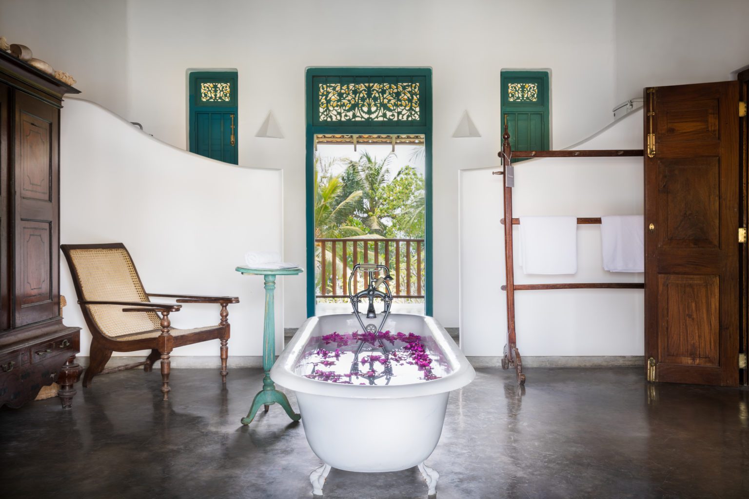 A bedroom with a claw-foot bath scattered with petals, a planter's chair and teal-framed doors to a balcony